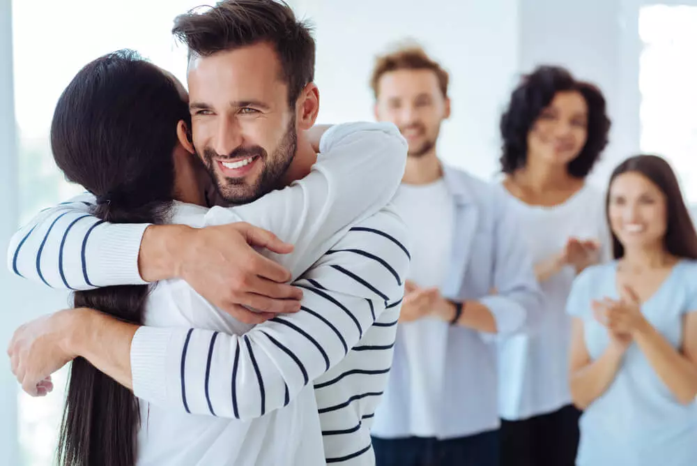 Couple hugging during rehab while a small support group applauds in the background, representing healing and connection in couples addiction treatment.