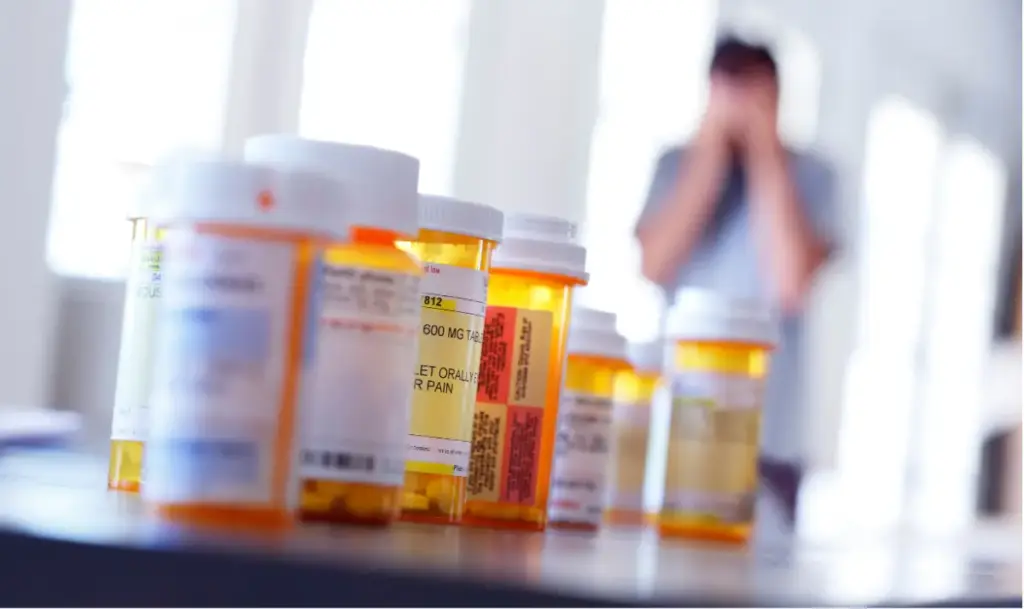 Multiple prescription opioid pill bottles in the foreground with a distressed person in the background representing opioid addiction and need for treatment